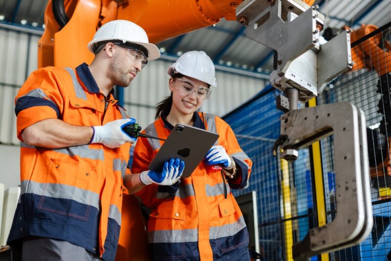 Dva inženjera u zaštitnoj opremi analiziraju industrijski sistem pomoću tableta pored robotske mašine