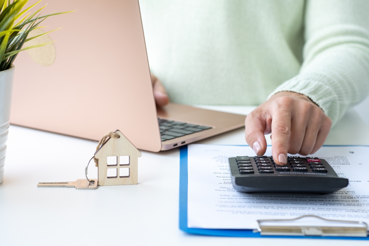 osoba koristi laptop i kalkulator, pored nje papiri i ključevi od kuće
